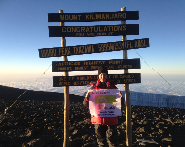 A person stands at Mount Kilimanjaro's summit sign, holding a fundraising banner for "Baraka Tanzania Porter Education Project," with clouds and rocky terrain below. Sign reads: "MOUNT KILIMANJARO CONGRATULATIONS YOU ARE NOW AT UHURU PEAK, TANZANIA, 5895 