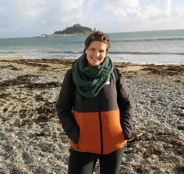 A person stands smiling on a pebbled beach, wearing an orange and black jacket with a green scarf, overlooking the sea and a distant island under a cloudy sky.