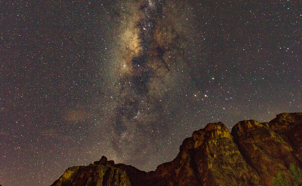 The Milky Way galaxy, bright and detailed, stretches vertically across a star-filled night sky above rocky, silhouetted cliffs.