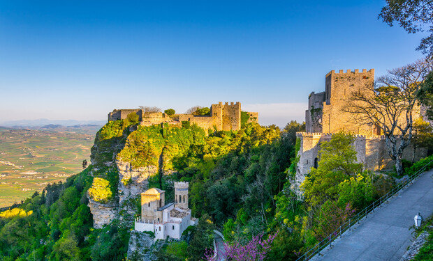 Ancient stone castle overlooks a lush, green valley, perched on a hilltop. A path winds along the edge, while trees surround the structure under a clear, blue sky.