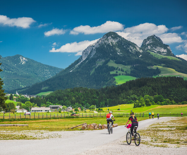 Cyclists ride on a gravel path through a lush green valley. Majestic mountains rise in the distance under a clear blue sky, while fields and a few buildings line the route.