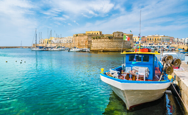 A small fishing boat rests docked in calm, clear waters beside an old stone fort surrounded by historic buildings and other boats in a sunny harbor setting.