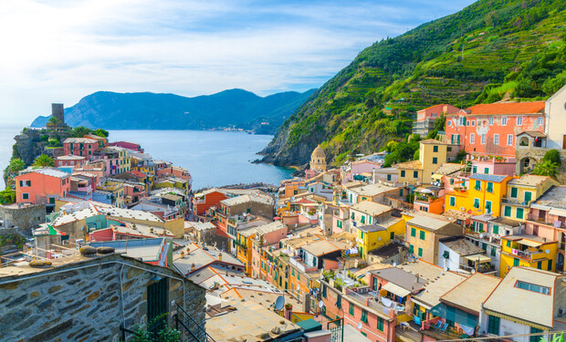 Colorful hillside buildings cascade down towards a calm blue sea, flanked by lush, green hills, under a clear sky. A distant tower stands on a rocky outcrop.