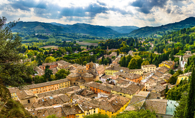 Historic town buildings with tiled roofs spread across a hilly landscape, surrounded by lush greenery and distant mountains, under a partly cloudy sky with sun rays peeking through.