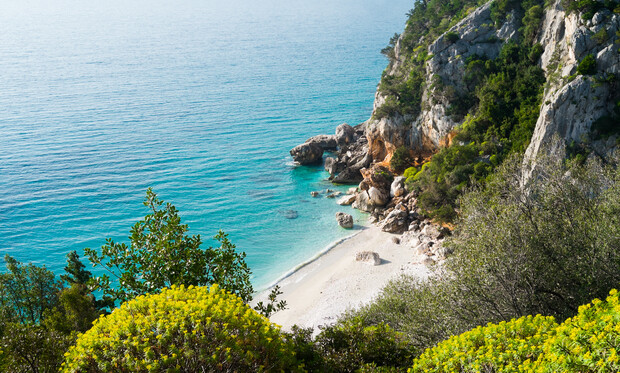 Turquoise water gently laps at a small, secluded sandy beach. Rocky cliffs and lush green foliage surround the tranquil cove, creating a peaceful and natural landscape.