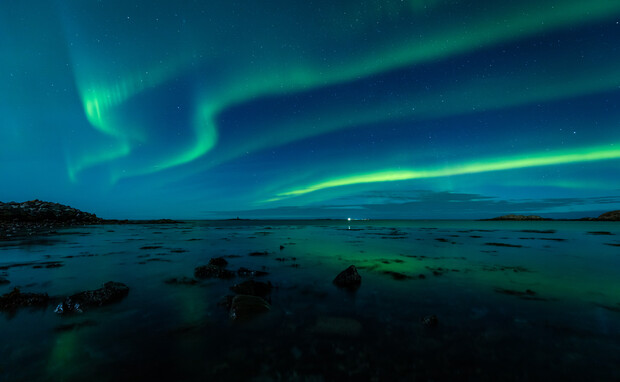 Green aurora borealis lights ripple across the night sky over a calm, rocky seascape, reflecting in the water. Distant landmasses and scattered clouds contribute to the tranquil scene.