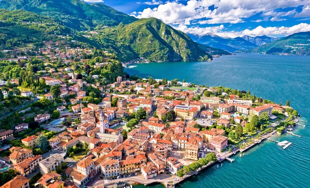 Red-roofed buildings cluster along a lakefront, surrounded by lush green hills under a blue sky with clouds. Boats are visible on the lake, enhancing the picturesque scenery.