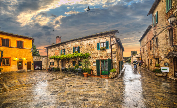 Stone house with green shutters sits quietly under a cloudy sky, surrounded by a wet cobblestone street in a quaint village setting, while a few birds fly overhead.
