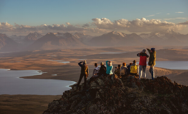 A group of people sits and stands on a rocky peak, observing a vast landscape of mountains and lakes under a cloudy sky, during golden hour, suggesting sunset or sunrise.