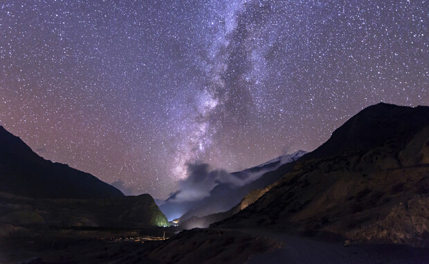 A vibrant Milky Way galaxy arcs across a star-filled sky. Below, rugged mountains and scattered lights dot a shadowed landscape, creating a serene nighttime wilderness scene.