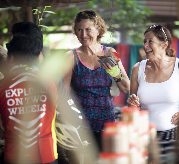 Two women, smiling and holding items, interact with a person wearing a cycling jersey labeled "EXPLORE! ON TWO WHEELS" in an outdoor setting with greenery and tables.