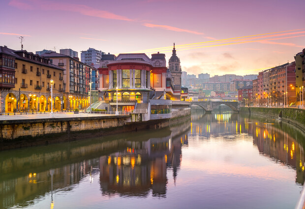 A large building with arched windows sits by a calm river at dusk. Surrounding are illuminated streets, historic architecture, and a distant bridge under a purple-orange sky.