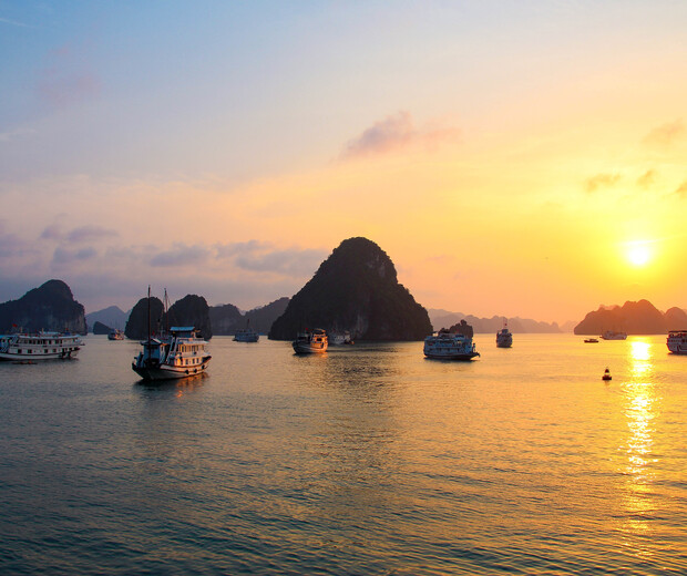 Boats float on calm water during a vibrant sunset, surrounded by rocky islands that create a scenic horizon, under a sky painted with warm orange and yellow hues.