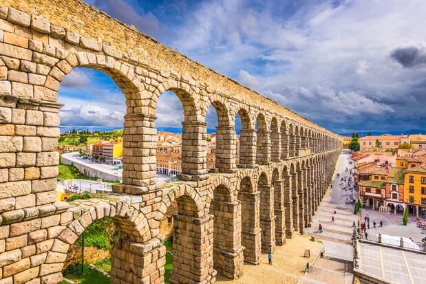 Ancient stone aqueduct with multiple arches spans a cityscape; people walk beneath it on a bright day, surrounded by colorful buildings and a dramatic cloudy sky.