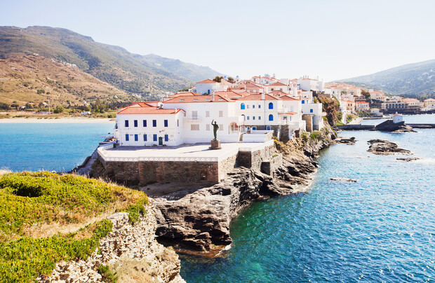 White coastal buildings with red roofs stand atop rocky cliffs by a vibrant blue sea. A statue overlooks the water, with mountains and more buildings in the distant background.