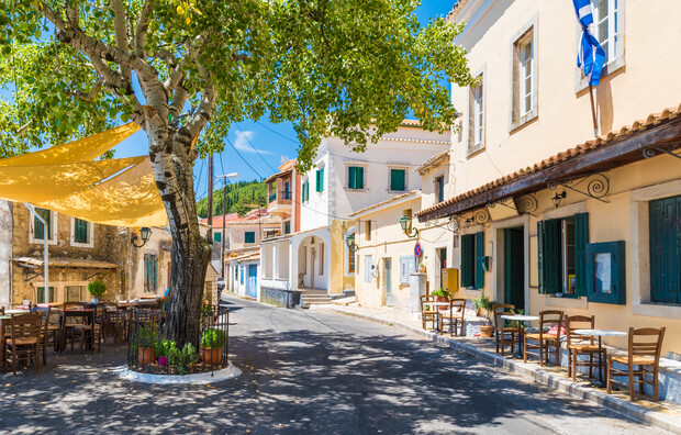Tree provides shade over tables and chairs on a sunny street lined with quaint, colorful buildings and vibrant greenery, creating a relaxed, picturesque village atmosphere.