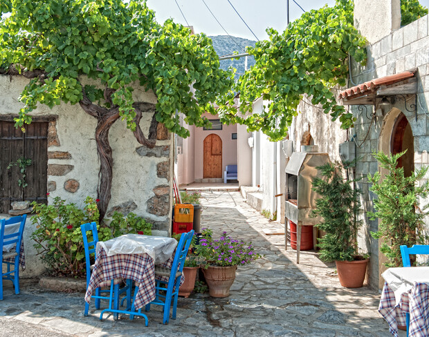 Café tables with checkered cloths sit under grapevine trellises, surrounded by rustic stone walls and potted plants, in a narrow, sunlit alleyway. A wooden door and mountains are visible in the distance.