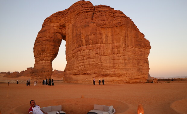 A large, naturally-formed rock arch towers under a clear sky. People stand and walk nearby on sandy terrain, with a seated man in foreground near outdoor furniture.