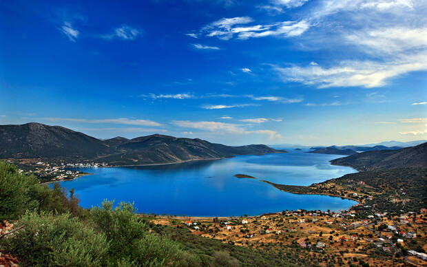 A tranquil blue lake stretches between rolling hills, with scattered houses along the shoreline under a clear, expansive sky. Lush greenery and dry grass frame the picturesque scene.