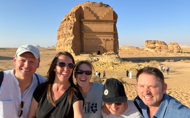 A group of five people smiling in front of a large, ancient rock-carved tomb in a desert setting, with other tourists and rocky formations visible in the background.