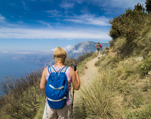 Two hikers walk along a narrow, winding trail on a coastal hillside. One carries a blue backpack and uses trekking poles, with the sea and distant mountains in the background.
