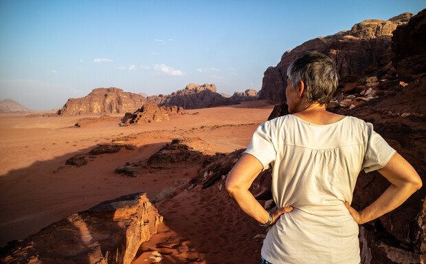 A person stands with hands on hips, looking across a vast desert landscape with red sand and rocky formations under a clear blue sky.