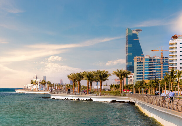 Coastal promenade lined with palm trees, adjacent to turquoise water. Skyscrapers and buildings rise in the background under a clear blue sky. People stroll along the pathway in a tranquil setting.