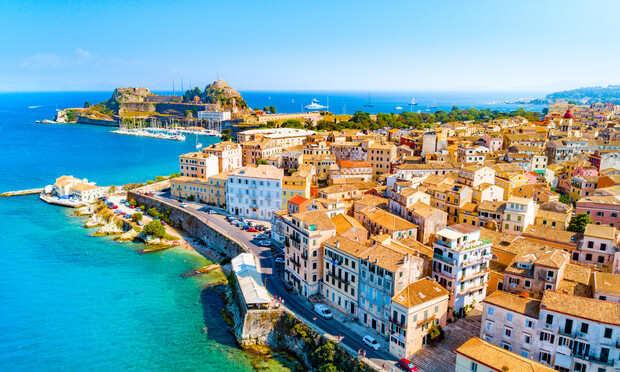Coastal town with terracotta-roofed buildings lining narrow streets; overlooks turquoise sea. A historical fort sits on a hill in the distance, and yachts are anchored in the bay.