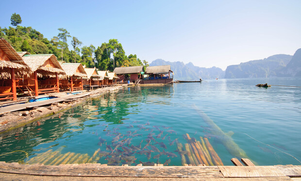 Thatched roof huts rest along a wooden dock by a clear, fish-filled, turquoise lake, surrounded by distant mountains and lush greenery under a bright, clear sky.