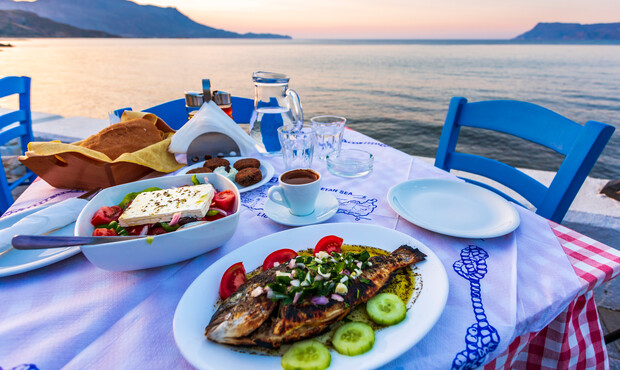 Grilled fish sits on a plate garnished with vegetables, accompanied by a Greek salad and bread on a table by the seaside at sunset, with blue chairs and calm water visible.