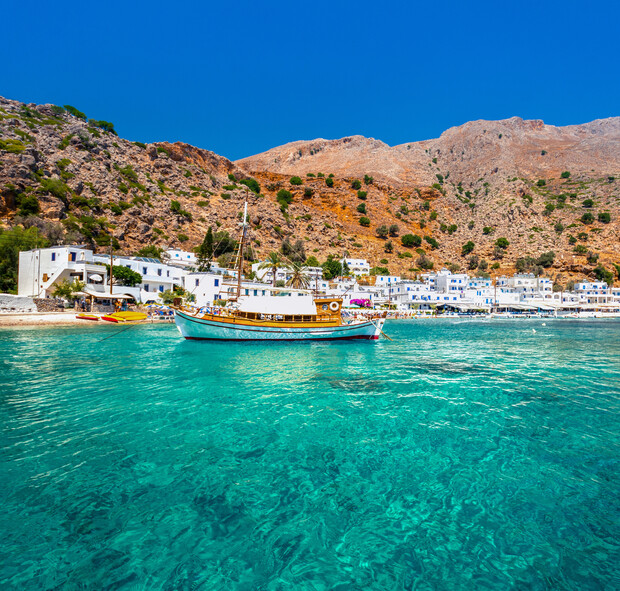 A wooden boat floats on clear turquoise water near a rocky coastline with white buildings and lush greenery under a bright blue sky.