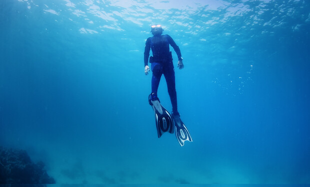 A scuba diver swims upward, wearing fins and a wetsuit, in a clear blue underwater environment with bubbles rising around.