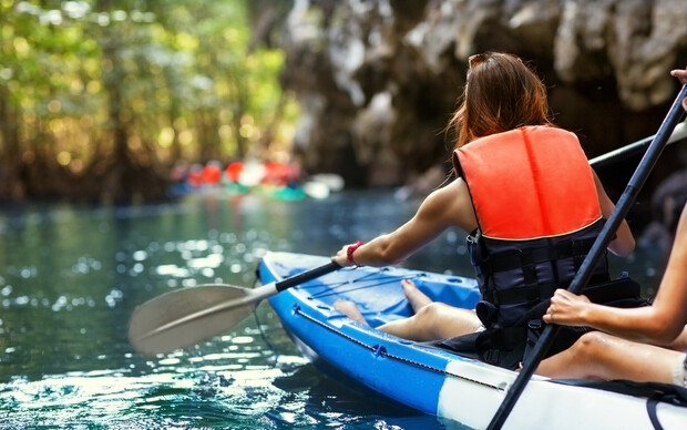 A person in an orange life vest paddles a kayak down a serene river surrounded by lush greenery and rocky cliffs. Other kayakers are visible in the distance.