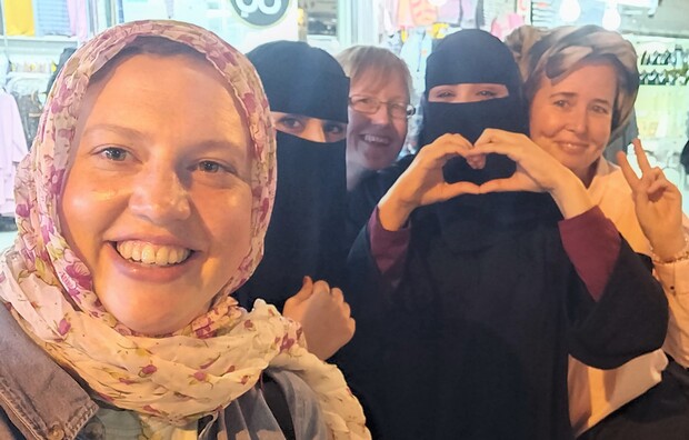 Five women smiling, one forming a heart with hands, another showing a peace sign. They are posing together in a vibrant, brightly lit market with clothing stalls in the background.
