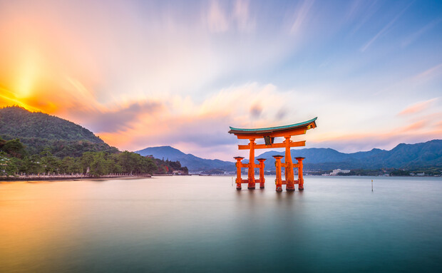 A large, traditional red torii gate stands in calm water, framed by a vibrant sunset. Mountains and a forested shoreline provide a serene backdrop, under a softly streaked sky.