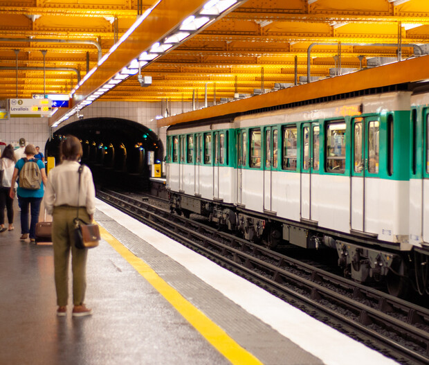 A green and white train is stationary on tracks as passengers walk on a platform under a brightly lit, yellow ceiling. A sign reads "Levallois."