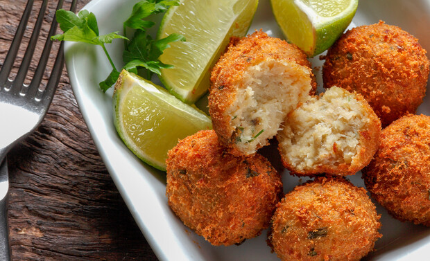 Golden fried balls, one split open, arranged in a white dish with lime wedges and parsley. A fork lies on a rustic wooden table nearby.
