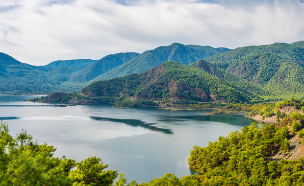 A calm lake reflects surrounding lush green mountains under a partly cloudy sky, creating a serene landscape. Dense forests cover the hillsides, enhancing the natural beauty of the scene.