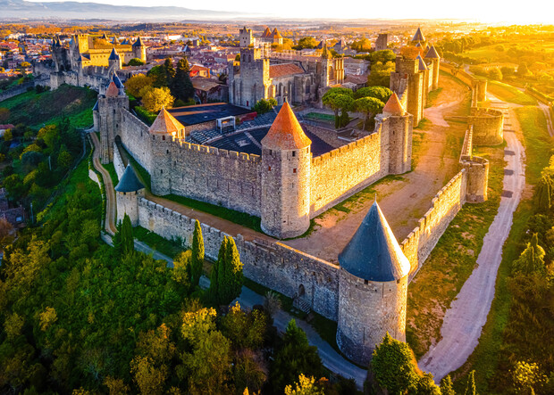 Stone castle with conical towers bathed in golden sunlight, surrounded by lush greenery and cityscape in the distance. The fortified walls dominate the landscape, exuding historical grandeur and architectural beauty.