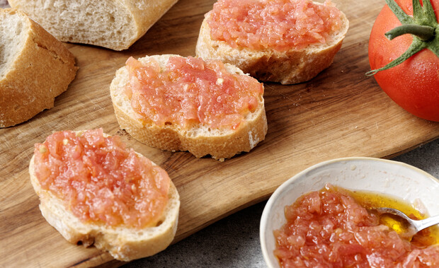 Slices of bread are topped with fresh tomato spread on a wooden board. A whole tomato and a bowl of extra spread with olive oil sit nearby.