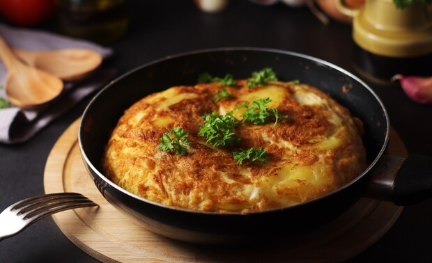 A cooked omelette rests in a frying pan, garnished with fresh parsley. The pan sits on a wooden board, with cutlery and cooking utensils nearby on a dark surface.