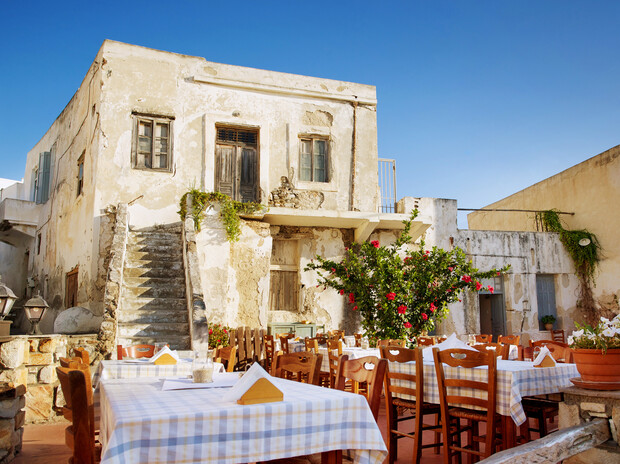 Weathered two-story building with blue shutters stands beside outdoor tables covered in checkered cloths. A flowering bush decorates the courtyard under a clear blue sky. No visible text.