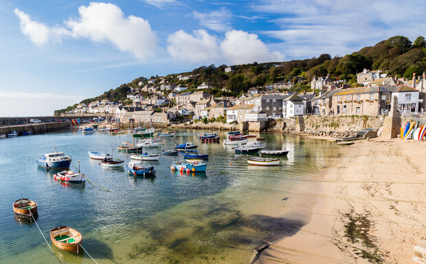 Boats float in a small, clear harbor along a sandy beach, with a quaint village of stone buildings and green hills in the background under a partly cloudy sky.