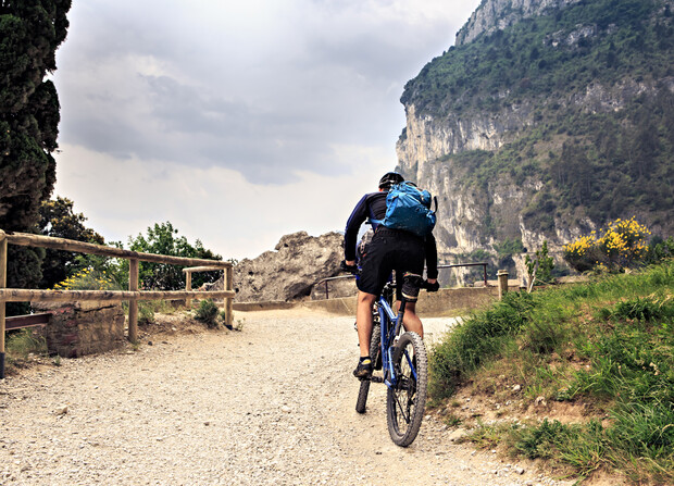 Cyclist rides a mountain bike uphill on a gravel path, surrounded by wooden fences, green vegetation, and steep cliffs under a cloudy sky.
