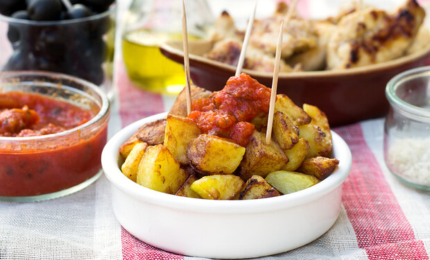 Golden roasted potatoes topped with red sauce and skewered by toothpicks rest in a white bowl on a checkered tablecloth, surrounded by olives and chicken dishes.