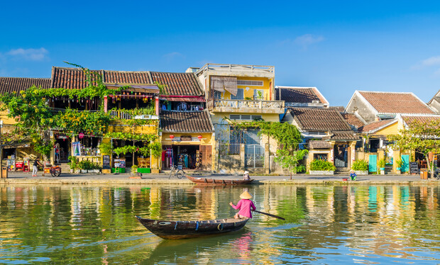 A person rows a small, wooden boat on a calm river, passing alongside a vibrant, historic town with colorful, vine-covered buildings beneath a clear blue sky.