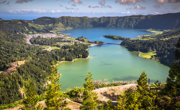 A serene lake lies within a volcanic crater, surrounded by lush green hills and a small village. The landscape features bright greenery, enhanced by scattered clouds in a clear sky.