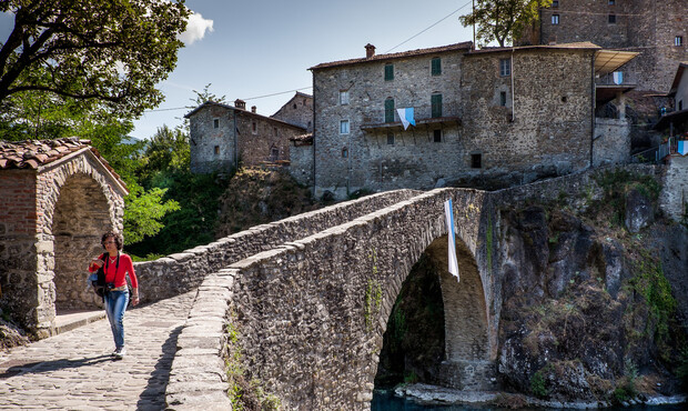 A person walks along a stone bridge with hanging laundry in a historic village. Stone buildings and greenery surround the scene, creating an old-world charm.