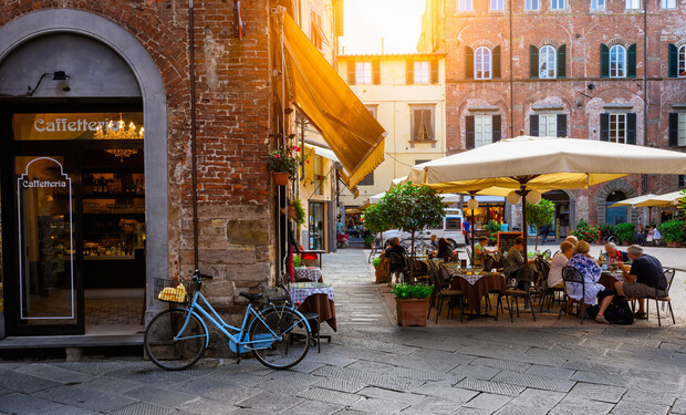 Bicycle leans against a brick wall outside a café named "Caffetteria," while diners sit at tables under umbrellas in a sunlit European square with historic buildings.