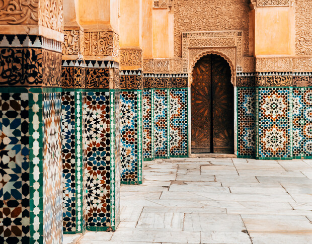 Ornate columns with colorful geometric mosaic tiles stand in a row, leading to a wooden door framed by intricate carvings, set within an elaborately decorated, stone-tiled courtyard.
